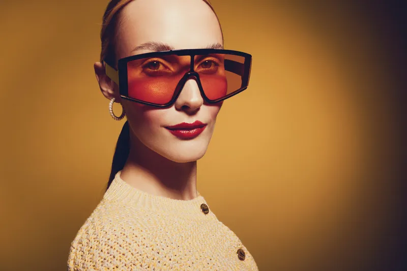 Stylish woman with oversized orange sunglasses and red lipstick poses confidently against warm yellow background. Fashion portrait with modern accessories and trendy makeup concept.