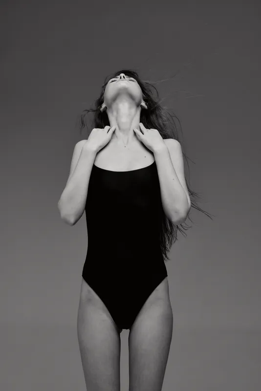 Black and white image of a woman in a black swimsuit with curly hair posing with head tilted back and hands near neck.