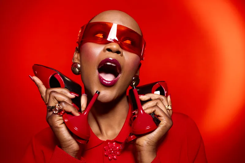 A confident Black woman with dark skin and bald head poses with red high heels and oversized red sunglasses against a vibrant red background. Fashion and bold style concept.