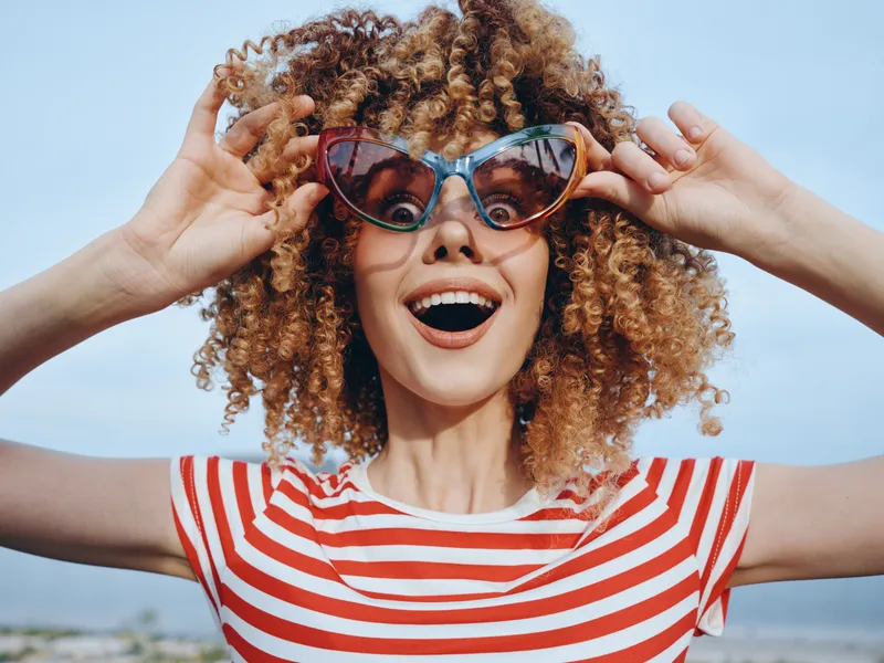 A joyful woman with curly hair wears oversized sunglasses and a red striped shirt, captured outdoors on a sunny day, exuding playful confidence and carefree vibe.