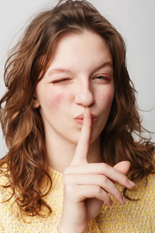 Young woman with wavy brown hair wearing yellow sweater makes shushing gesture with finger on lips and winks. Concept of silence, secret, quiet, and communication.