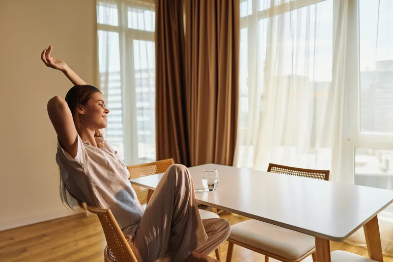 Woman stretching in morning at home by dining table with sunlight through curtains. Relaxed casual female sitting on chair, enjoying peaceful bright interior and warm cozy atmosphere.