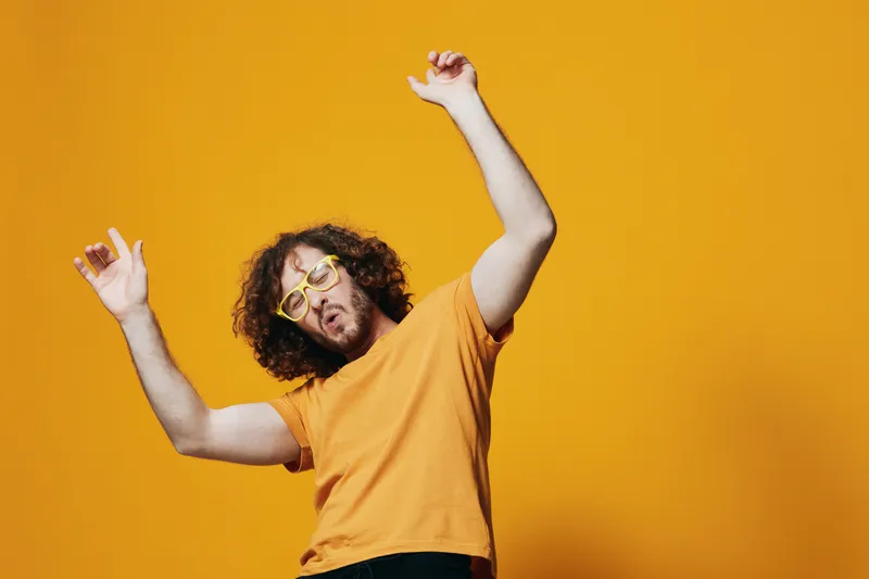 Young man dancing joyfully against a vibrant yellow background, wearing quirky yellow glasses and a casual orange t shirt, radiating energy and positivity