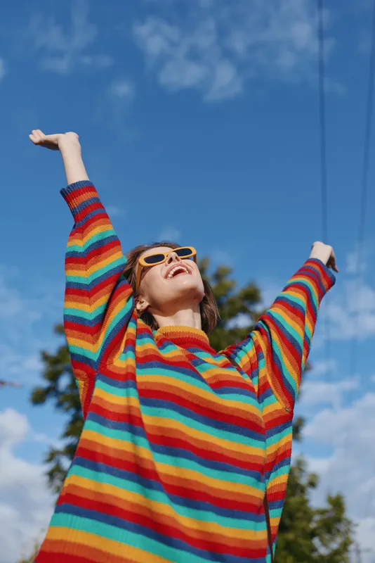 Woman in rainbow sweater with sunglasses and arms raised, happy and joyful portrait outdoors under blue sky, colorful knitwear and carefree expression during a sunny day in park