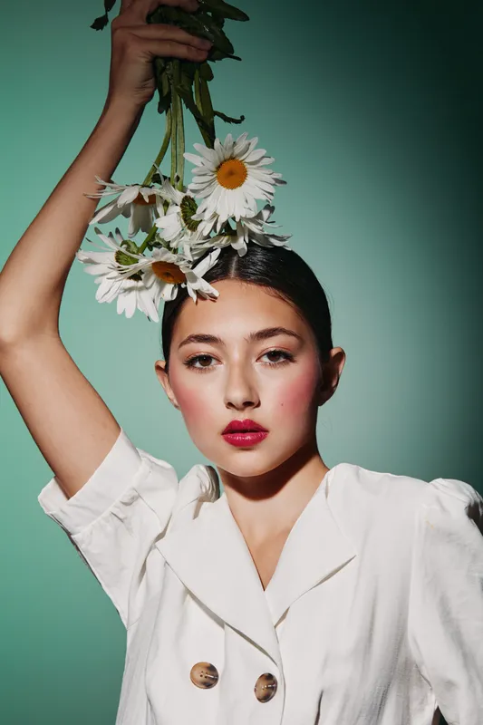 Young woman with sleek hair holding white daisies above head wearing white fashion jacket. Bold pink lips and delicate makeup on soft green studio backdrop. Beauty creative concept.