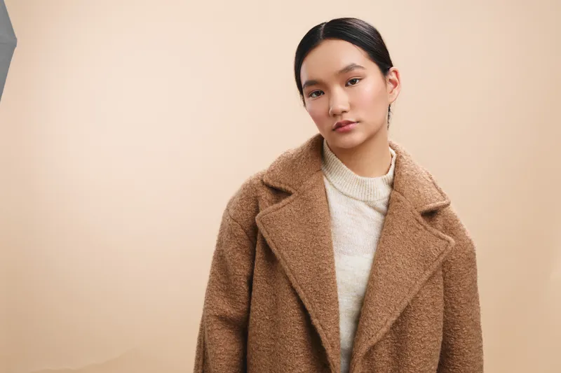 Asian woman wearing a warm coat stands against a soft backdrop, presenting a calm, stylish look. She gazes away, conveying poise and thoughtful mood in a contemporary portrait.