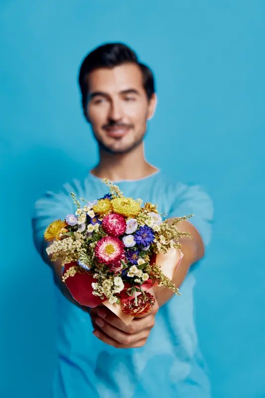Young smiling man holding a colorful bouquet of flowers against a bright blue background, showcasing joy and appreciation in a cheerful lifestyle concept.