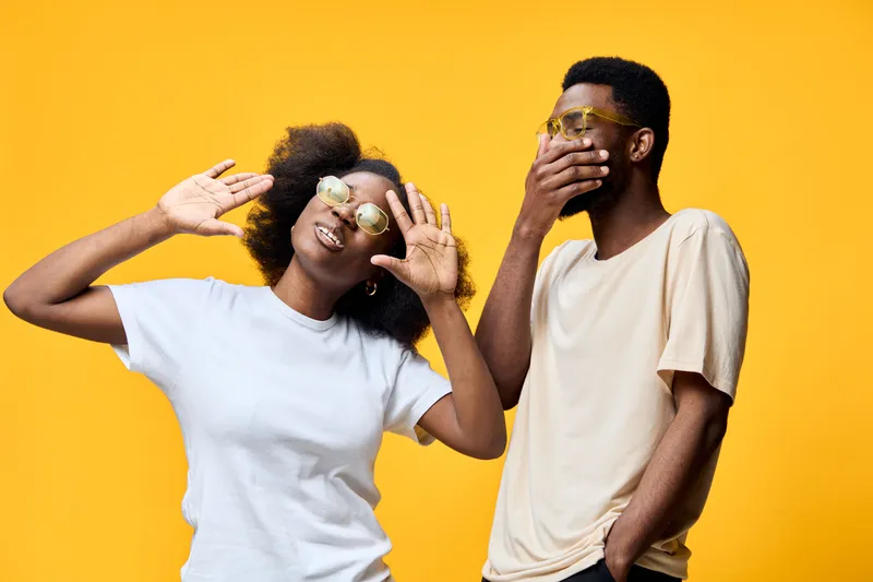 Joyful young Black couple enjoying a playful moment with bright yellow background, wearing stylish casual clothing and sunglasses