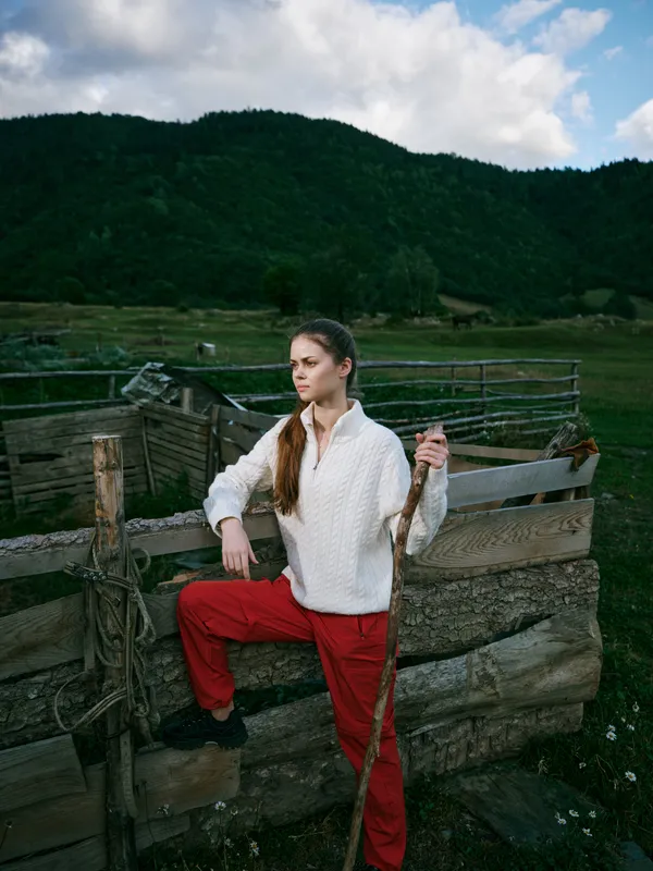 Woman on a rustic fence in a green countryside, holding a wooden staff, wearing white sweater and red pants, with hills in the background and cloudy sky.
