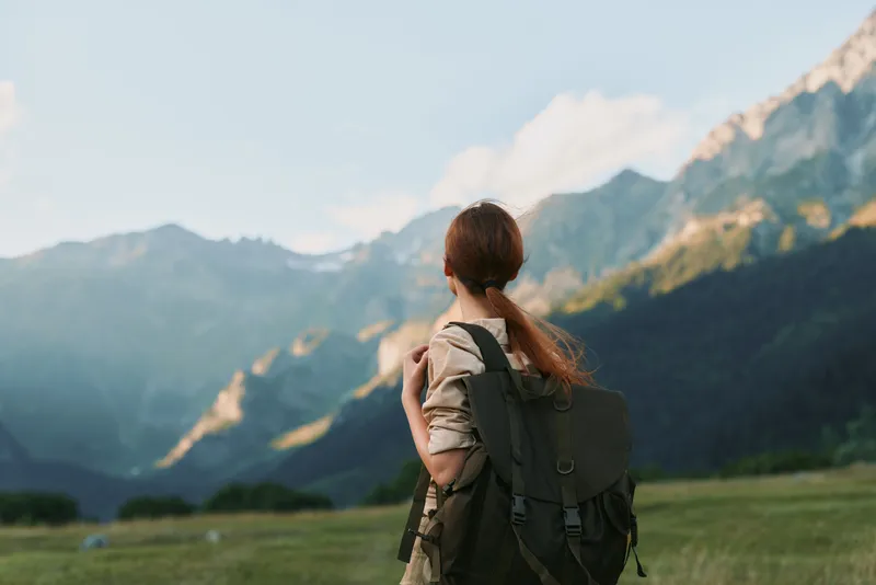 Young woman with a ponytail carries a large backpack while hiking in a mountain valley under a clear sky during daytime.