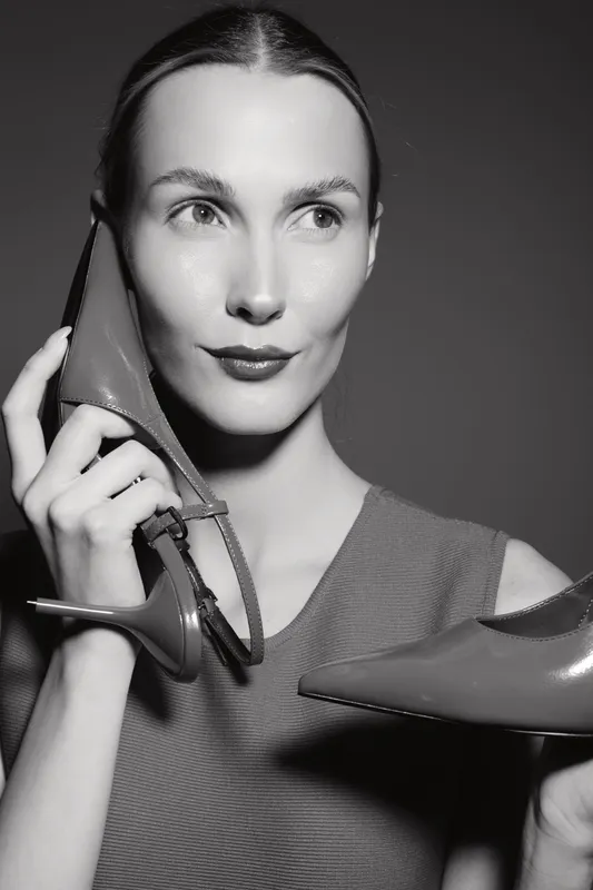 Young white woman with fair skin holds red high heel shoes near her face in black and white studio portrait concept of fashion and elegance.