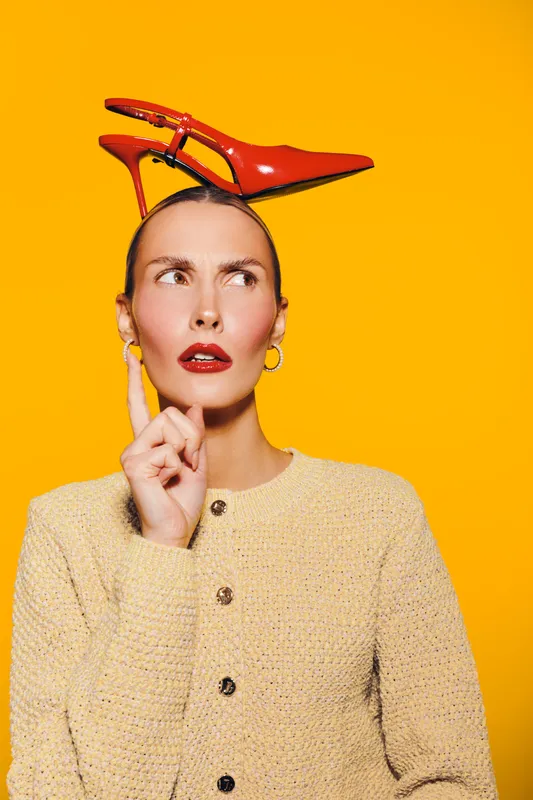 Thoughtful woman with red high heel shoe balanced on head, wearing beige cardigan and gold hoop earrings. Bright yellow background creates vibrant contrast. Concept for fashion, creativity, and style.