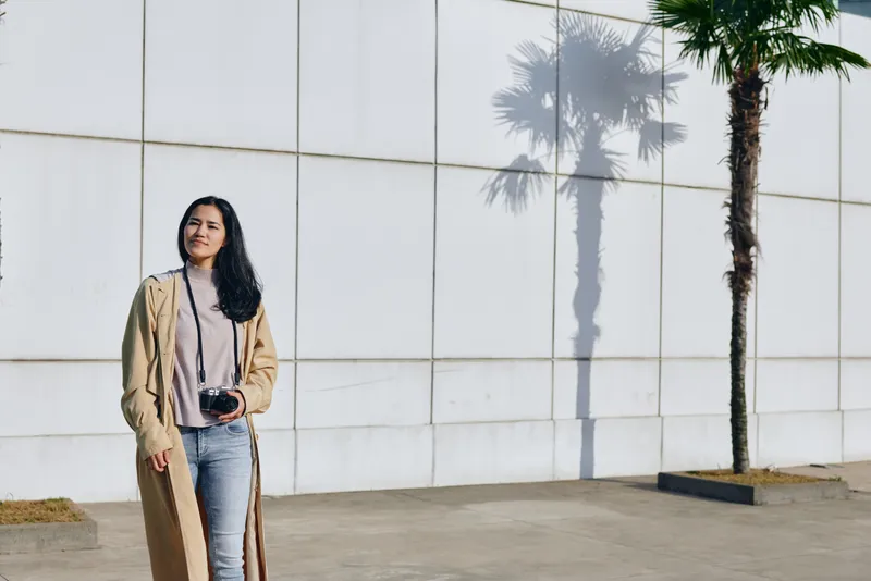 Woman with camera in casual coat stands outdoors beside a white wall, showcasing fashion, street style, and urban mood for lifestyle photography and visual storytelling