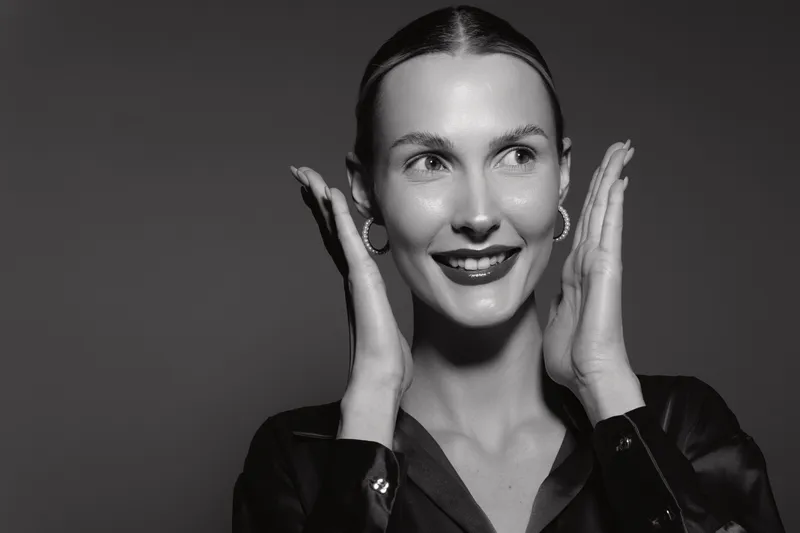 Elegant Caucasian woman with fair skin and dark blouse poses with raised hands, smiling and looking away in black and white studio portrait.