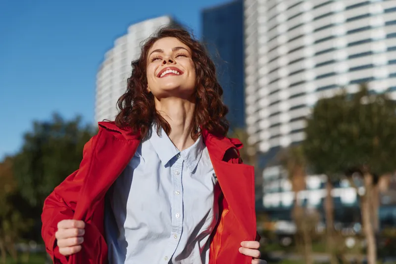 Young woman with curly hair enjoys a sunny day outdoors wearing a red jacket and blue shirt with a joyful smile and closed eyes.