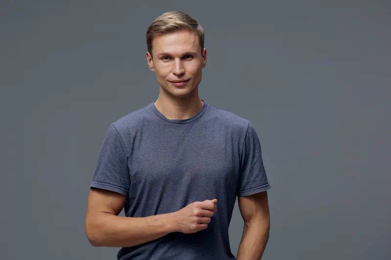 Confident young man with fair hair wearing a casual blue t-shirt posing against a plain gray background in a studio. Happy, relaxed, smiling, male, casual style, single person, portrait, lifestyle, in