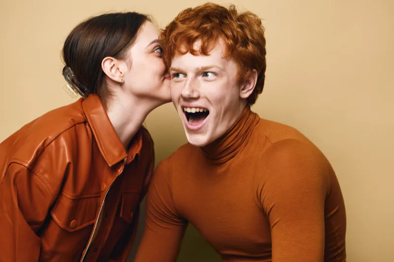 Young woman whispering secret to surprised young man in casual brown clothing on beige background, friendship and playful moment captured with emotions and expressions.