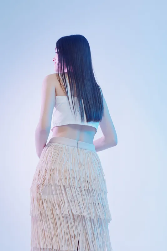 Young woman with long dark hair wearing a white top and beige fringed skirt stands against a light background. Concept of beauty fashion style and elegance in studio lighting.