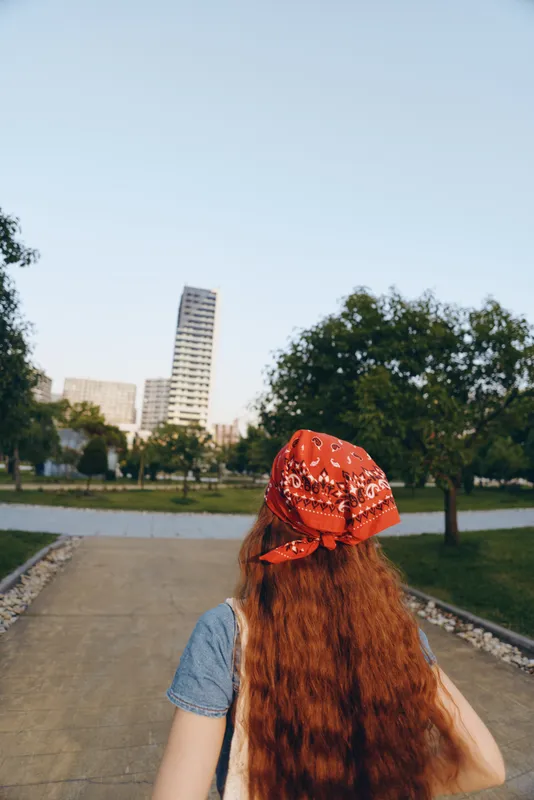 Lifestyle woman with long red hair in modern boho-western denim street style wearing a red bandana, walking outdoors with urban buildings and trees in soft film color.