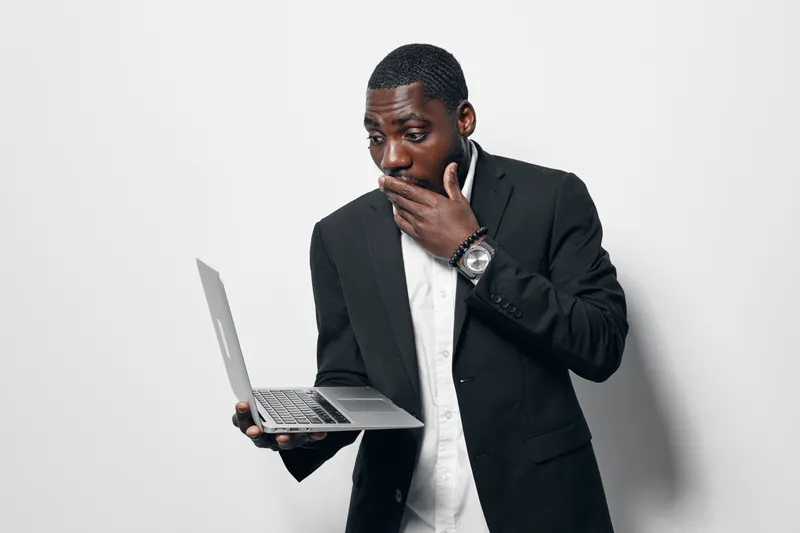 Black man with dark skin in formal suit holding laptop and looking surprised with hand on mouth against white background. Business, technology and emotion concept.
