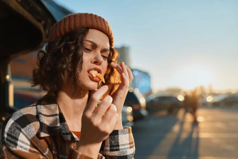 woman snack car beanie outdoors sunset candid emotion: Young woman wearing a beanie eats a snack by a car trunk outdoors at sunset, plaid jacket, golden hour glow, authenticity and mindful living.