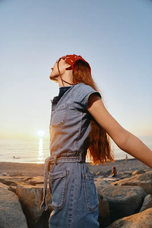 Lifestyle woman in modern boho-western denim street style enjoying sunset outdoors, relaxed and free-spirited with film color warmth on rocky beach background.