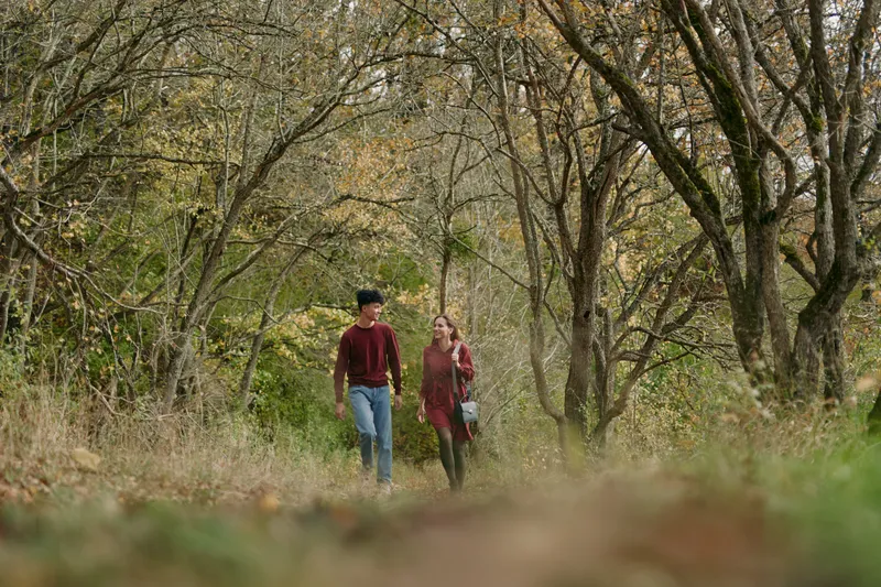 A young couple walking along a quiet forest path in autumn, sharing a genuine moment of connection and conversation. Natural light and candid composition emphasize authenticity and credible emotion.