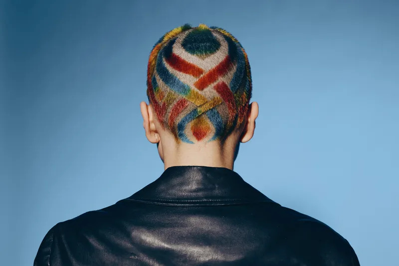 woman with colorful shaved hair design wearing black leather jacket against blue background, creative hairstyle, modern fashion, artistic expression, unique look