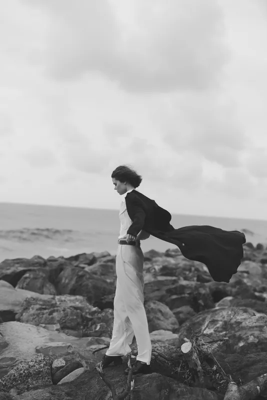 Coastal wind cape dress silhouette on moody rocks by the ocean, monochrome movement captures dramatic fashion and solitary elegance against a windy shoreline