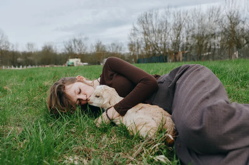 A woman lies on grassy ground in a field, embracing a young goat kid, calm and content. A lifestyle moment of bonding with animals and nature, highlighting tranquility, companionship, and simple