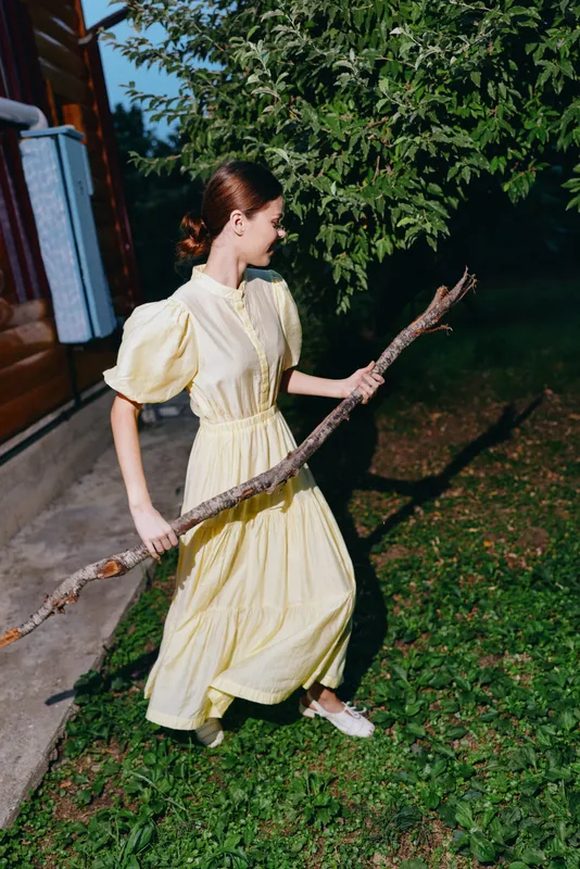 Woman in a yellow dress walks through a garden lawn, holding a long branch, enjoying outdoor light, soft shadows, green surroundings, and a tranquil natural atmosphere.