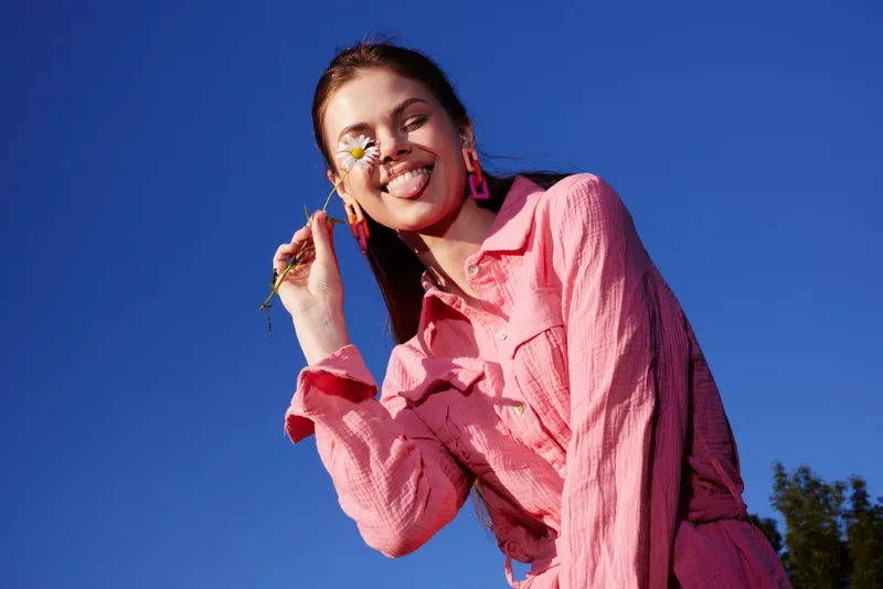 Cheerful woman in pink clothing holding a flower, smiling against a bright blue sky, showcasing happiness and warmth in a vibrant outdoor setting