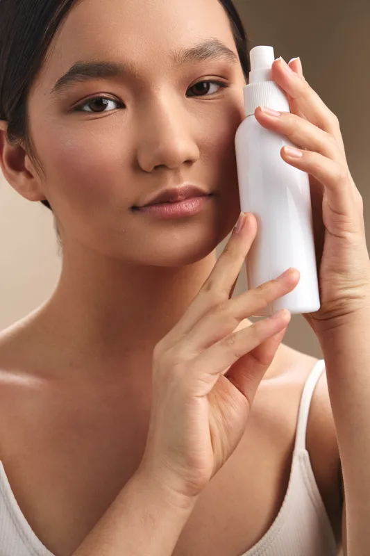 asian woman holds a white bottle near her face in a close up portrait, applying skincare with a gentle touch, calm expression, soft lighting, and a clean modern beauty routine vibe.