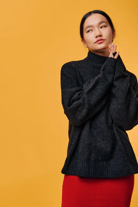 Young woman wearing black sweater and red skirt posing thoughtfully with hands near face against orange background. Female model with contemplative expression and casual clothing style.
