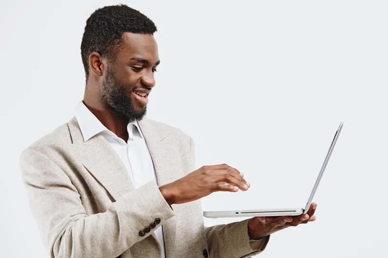 Smiling man with dark skin, wearing beige blazer and white shirt, holding laptop and looking at screen, studio portrait with plain white background, casual professional appearance. People lifestyle co