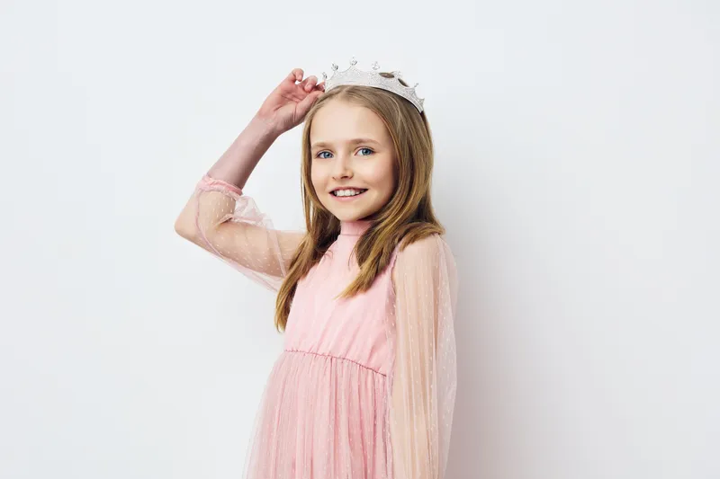 Joyful little girl wearing a pink dress and a crown with a playful smile against a light background, capturing a moment of youthful innocence and happiness