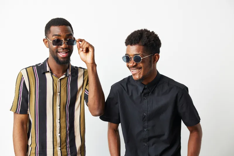 Two cheerful black men enjoying a fun moment, wearing stylish sunglasses. Their vibrant shirts reflect a joyful and relaxed atmosphere in a minimalist setting.