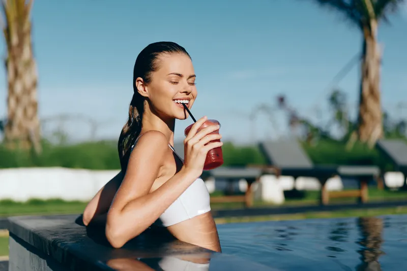 Young woman by the pool edge enjoys a refreshing beverage, smiling in a white bikini, capturing a sunny outdoor summer lifestyle moment of relaxation and leisure