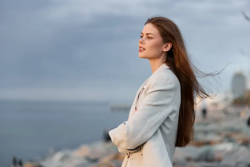 Confident businesswoman by the sea, showcasing elegance and strength with flowing hair against a dramatic coastal background.