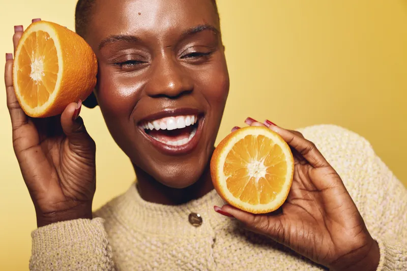 Happy African woman with dark skin holds two orange halves near face, smiling with closed eyes. Concept of healthy lifestyle, fresh fruit, vitamin C, and natural beauty.