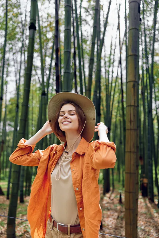 woman bamboo forest hat orange smile nature portrait of a young traveler wearing casual jacket, enjoying sunlight and serene scenery while strolling among tall green trees in outdoor adventure
