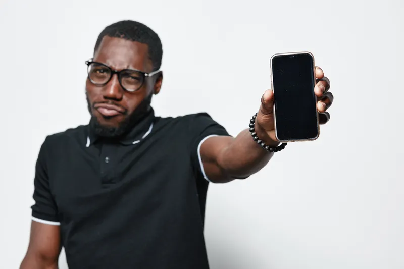 African American man with dark skin wearing glasses and black polo shirt showing smartphone with blank screen in hand isolated on white background. Technology, communication and casual style.
