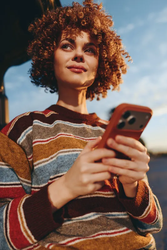 Woman with curly hair outdoors, smiling gently while holding a smartphone, striped sweater for woman lifestyle, warm sunset glow, relaxed posture, soft wind, open sky background sunlit