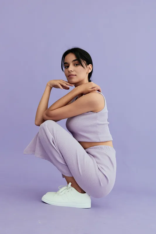 Young woman in lilac knitwear crouching against a matching purple background, wearing white sneakers and displaying a calm, thoughtful expression.