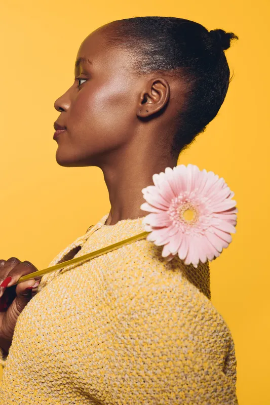 Profile of a young black woman with dark skin holding a pink flower behind her shoulder, wearing a yellow sweater. Studio portrait on yellow background with soft lighting and natural beauty concept.
