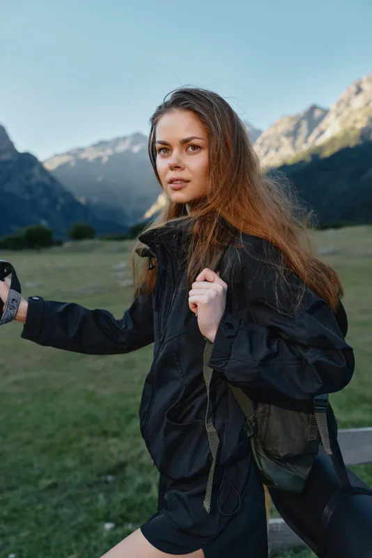 A young woman in a black jacket stands in a valley with rugged mountains behind her. She faces the camera with a gentle smile, creating a confident, vibrant outdoor portrait.