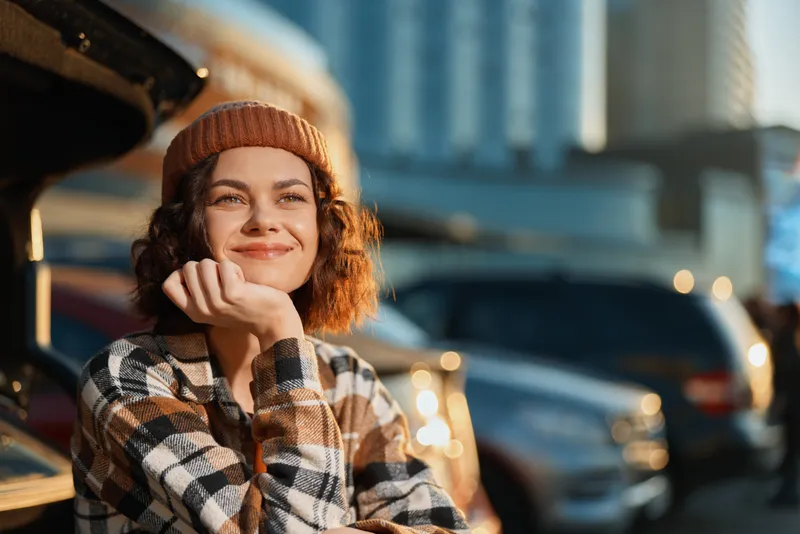 Woman smile beanie urban candid golden sunlight plaid shirt portrait in parking lot, authenticity and candid lifestyle moment with emotional storytelling and mindful living vibe.
