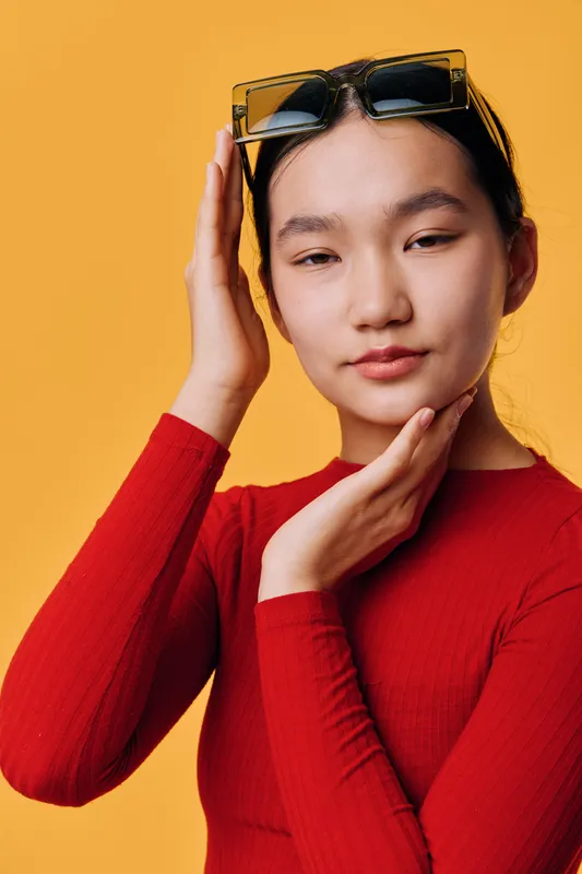 Young woman in red shirt posing with sunglasses on head and hand touching face against yellow background. Fashion, beauty, portrait, casual style with confident expression and natural makeup.