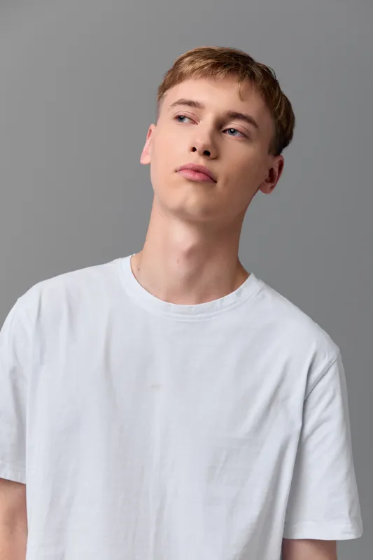 Young man in a white t shirt stands in a neutral studio, looking off to the side with a thoughtful expression. Clean light and minimal background emphasize casual style and quiet confidence
