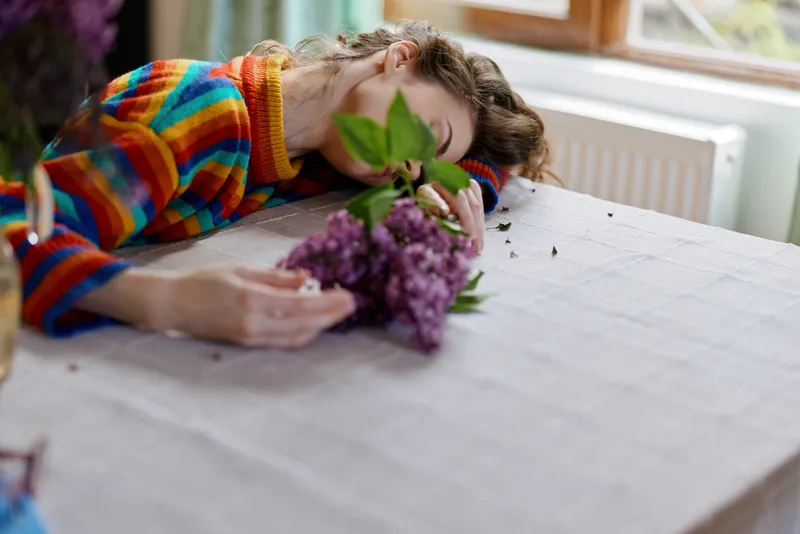 Young woman lies across a bright indoor table, smelling a fresh bouquet with eyes closed, a peaceful intimate moment